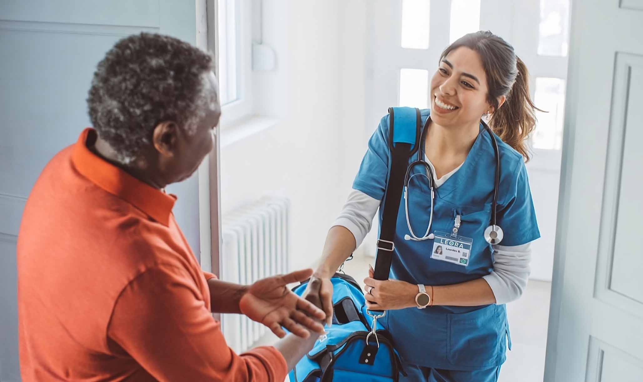 A leora home health aide coming to a patients door wearing blue scrubs and shaking patients hand